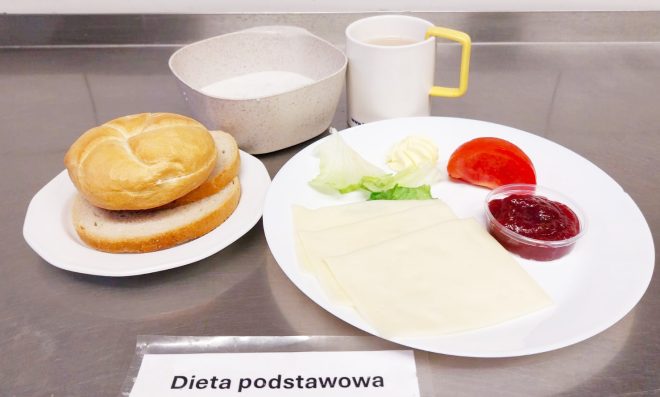 Na talerzu leży pszenna bułka i dwie kromki chleba. Obok ułożono dwa plasterki sera żółtego, liść sałaty, kawałek pomidora, porcję masła oraz niewielki pojemniczek z dżemem. W tle znajduje się miseczka z białym mlecznym napojem oraz kubek z kawą z mlekiem. Całość opisana jako „Dieta podstawowa”