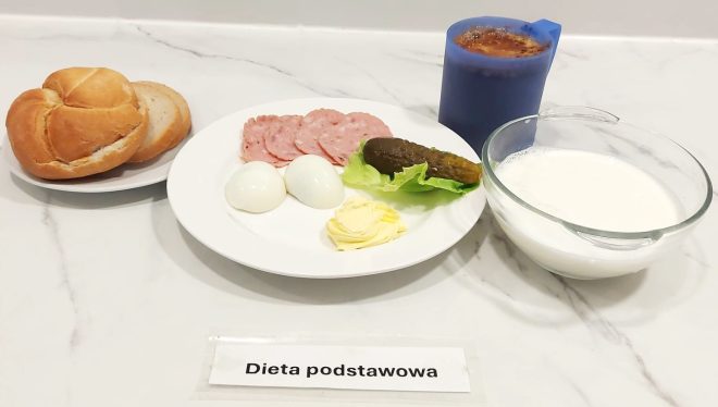 Śniadanie: na talerzu plasterki kiełbasy, dwa jajka gotowane na twardo, masło, ogórek kiszony i liść sałaty. Obok pieczywo pszenne (bułka kajzerka i kromki chleba). W tle niebieski kubek z kawą z mlekiem oraz miseczka z mlekiem. Na dole napis „Dieta podstawowa”