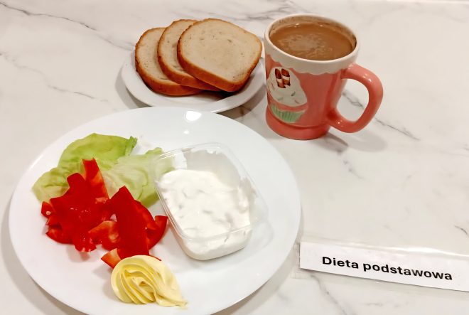 „Dieta podstawowa” Śniadanie na białym talerzu: liście sałaty, plastry czerwonej papryki, porcja twarożku i masło, obok trzy kromki chleba pszennego na osobnym talerzyku oraz kubek kawy z mlekiem w różowym kubku z motywem babeczki