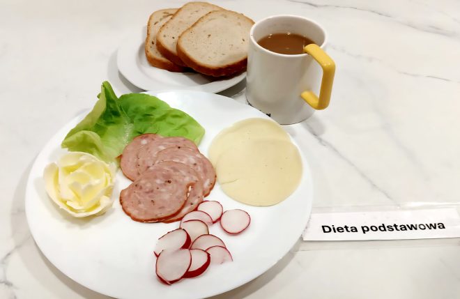 Talerz z liściem sałaty, masłem uformowanym w kształt róży, plastrami kiełbasy, plastrami sera żółtego oraz plasterkami rzodkiewki. W tle talerzyk z trzema kromkami chleba pszennego i kubek kawy z mlekiem. Posiłek w diecie podstawowej