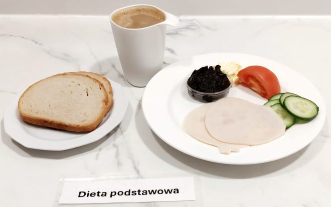 Śniadanie w diecie podstawowej – na talerzu bułka kajzerka i kromki chleba, obok kawa z mlekiem, talerz z plastrami wędliny drobiowej, masłem, cząstką pomidora, plastrami ogórka i porcją sałatki z buraczków; w miseczce biała zupa mleczna