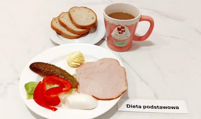 Porcja pieczywa pszennego podana z plastrem gotowanej wędliny, ogórkiem kiszonym, kawałkiem czerwonej papryki, liściem sałaty, ćwiartką jajka na twardo i porcją masła. Do zestawu filiżanka kawy z mlekiem w ozdobnym kubku