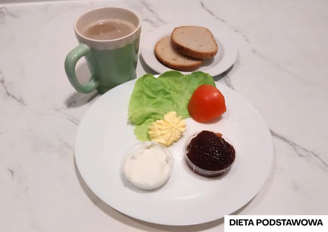 Na talerzu śniadaniowym w ramach „Diety podstawowej” podano liść sałaty, pomidora, porcję masła, porcję twarożku i porcję dżemu. Obok stoją dwie kromki chleba pszennego oraz kubek kawy z mlekiem