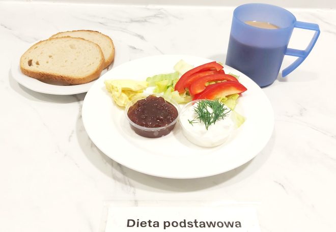 Śniadanie w diecie podstawowej: na talerzu znajdują się plasterki czerwonej papryki i sałata, porcja twarożku ze świeżym koperkiem, masło oraz mały pojemniczek z dżemem. Do zestawu podano dwie kromki chleba pszennego oraz kawę z mlekiem w niebieskim kubku