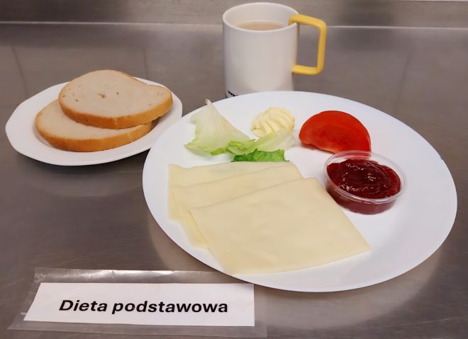 Śniadanie w diecie podstawowej – talerz z trzema plastrami żółtego sera, liśćmi sałaty, kawałkiem pomidora, porcją masła i dżemem. Obok dwie kromki chleba, kubek z kawą z mlekiem. Podpis: dieta podstawowa
