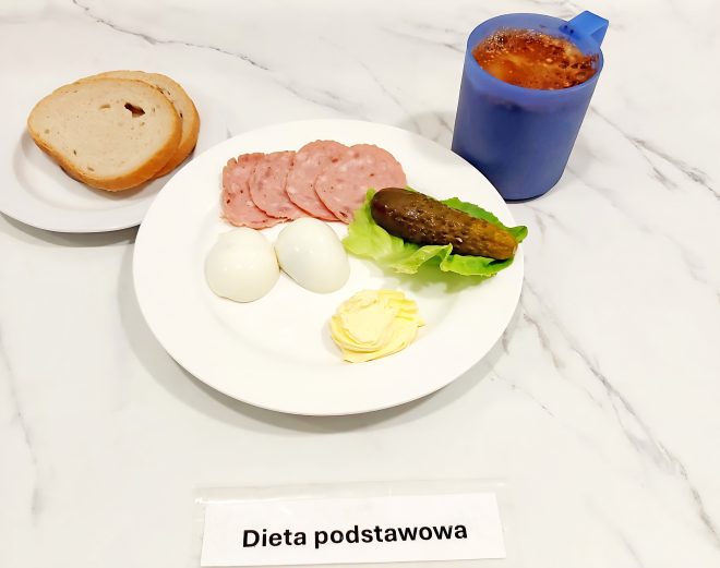 Na talerzu podano dwa jajka ugotowane na twardo, kilka plasterków wędliny, porcję masła oraz ogórka kiszonego na liściu sałaty. Obok znajduje się talerzyk z kromką chleba i bułką pszenną. Do zestawu dołączono napój w niebieskim kubku. Całość opisana jako „Dieta podstawowa”