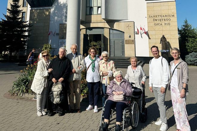 Grupa seniorów i opiekunów pozuje do wspólnego zdjęcia przed wejściem do Sanktuarium św. Ojca Pio w Warszawie, obok widoczny pomnik świętego