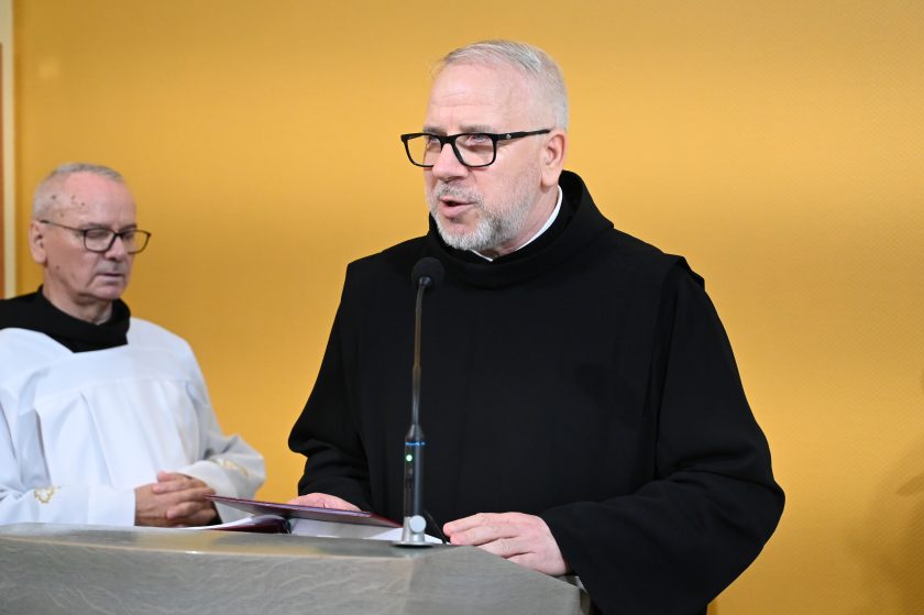 Zakonnik w czarnym habicie i okularach odczytuje tekst przy ambonie. Obok niego stoi starszy duchowny w białej albie, z dłońmi splecionymi w geście modlitwy