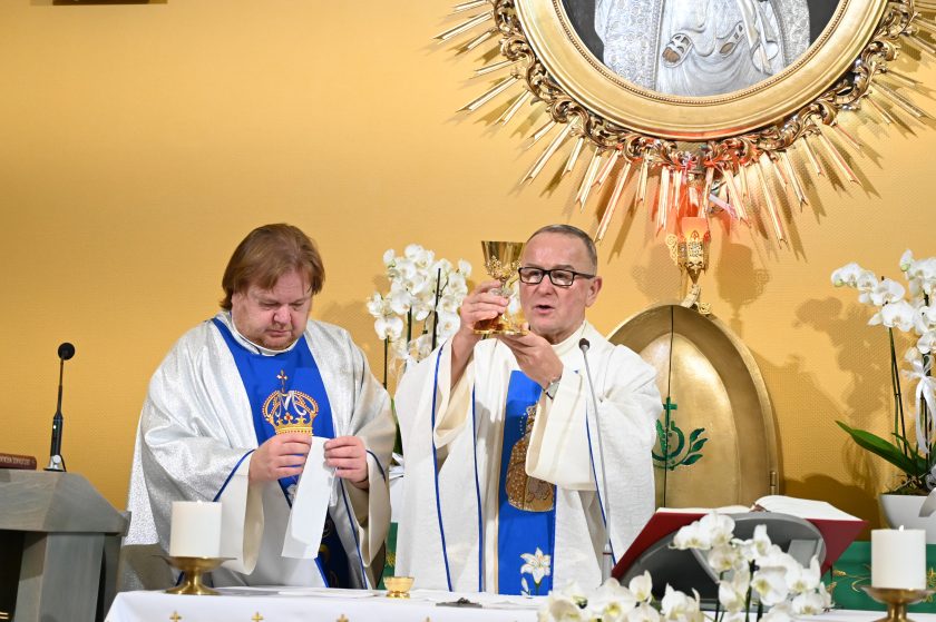 Dwóch kapłanów podczas Mszy Świętej przy ołtarzu – jeden z nich unosi złoty kielich eucharystyczny, a drugi przygotowuje liturgiczne naczynia. W tle obraz Matki Bożej w złotej ramie otoczonej promieniami oraz białe kwiaty.