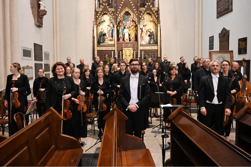 Orkiestra i chór ustawione w nawie bocznej kościoła: smyczki siedzą z instrumentami, za nimi stoją chórzyści w czerni z teczkami nut. Na środku tyłem do kamery dyrygent w fraku; przed nim siedzący muzyk z batutą na kolanach i mikrofonem na statywie. W głębi ołtarz tryptyk ze złoceniami; ogólna gotowość do rozpoczęcia utworu.