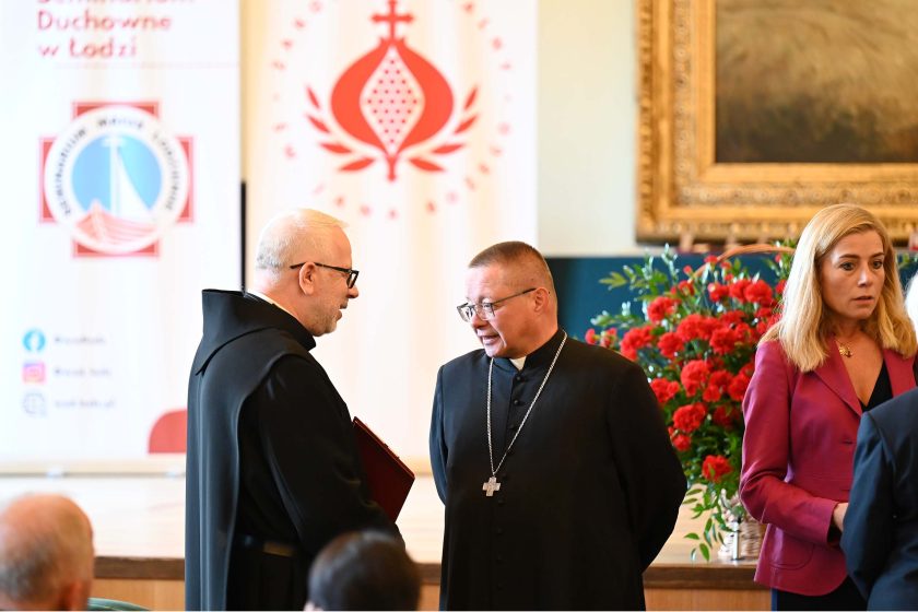 Wnętrze sali uroczystości. Po lewej zakonnik w czarnym habicie i okularach trzyma bordową teczkę, rozmawia z duchownym w czarnej sutannie z długim łańcuchem i krzyżem pektoralnym. Po prawej kobieta w malinowej marynarce spogląda w bok. Za postaciami kwitnąca kompozycja czerwonych goździków i roll-upy z emblematem zakonu; pierwszy plan miękko rozmyty.