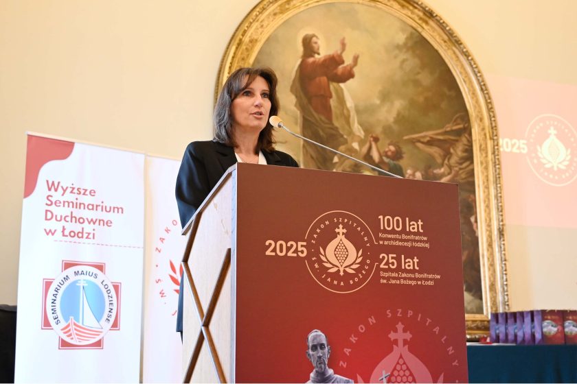 Kobieta z ciemnymi, półdługimi włosami przemawia z mównicy z bordowym frontem i znakami jubileuszu „2025 • 100 lat… • 25 lat…”. Po lewej stoją dwa roll-upy (m.in. „Wyższe Seminarium Duchowne w Łodzi”), w głębi duży, owalny obraz w złotej ramie z postacią w czerwonej szacie unoszącą dłonie. Na prawo fragment ekranu z projekcją logo jubileuszu.