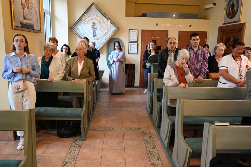 Wierni stojący w ławkach kaplicy w czasie nabożeństwa. W centrum zakonnica w szarym habicie, a obok starsze kobiety i osoby w różnym wieku. Nad wejściem widoczny napis „KONFESJONAŁ”
