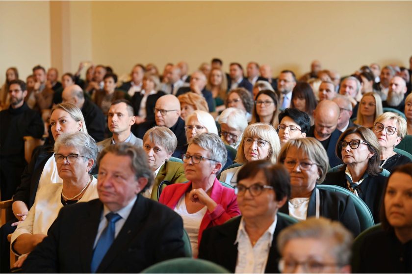 Szerokie ujęcie publiczności siedzącej w rzędach zielonych, tapicerowanych foteli. W pierwszych rzędach przeważają kobiety w okularach; twarze skupione, część osób lekko uśmiechnięta, część notuje lub patrzy na scenę. Tło wypełniają kolejne rzędy słuchaczy w różnym wieku, oświetlenie ciepłe, dzienne.