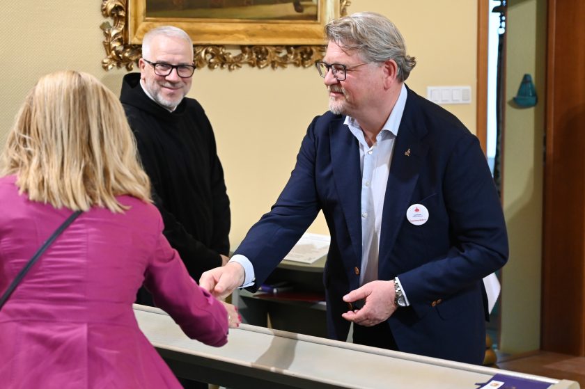 Mężczyzna w granatowej marynarce wita się i podaje dłoń kobiecie w różowym żakiecie. Obok zakonnik w czarnym habicie uśmiecha się, obserwując tę scenę