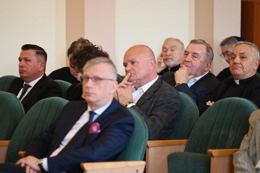 Ujęcie boczne fragmentu widowni. Kilku mężczyzn w garniturach siedzi w zielonych fotelach, większość z poważnym wyrazem twarzy; jeden z dłonią przy ustach w geście zamyślenia. W dalszym planie starsi uczestnicy i duchowny w koloratce; światło miękkie, ściany jasne.