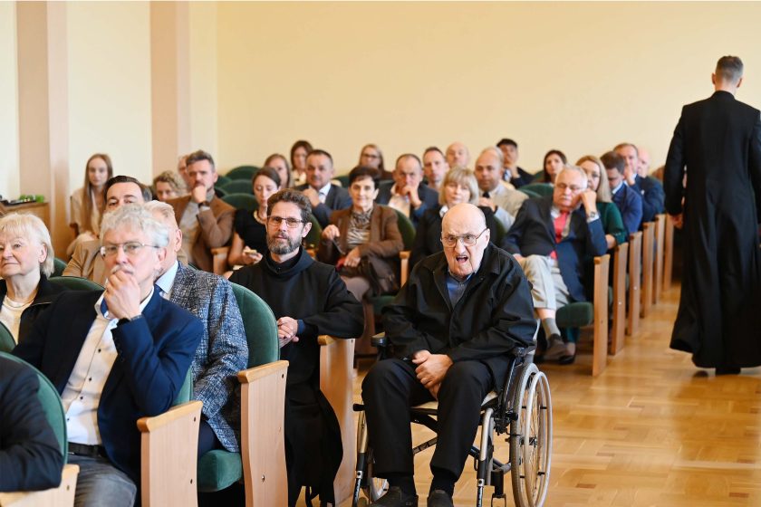 Widownia w głębi sali; na pierwszym planie starszy mężczyzna na wózku inwalidzkim, obok niego zakonnik w czarnym habicie. Wokół rzędy słuchaczy, część patrzy w stronę mównicy, część rozmawia szeptem. Po prawej w przejściu widać sylwetkę duchownego odchodzącego w kierunku wyjścia; podłoga z jasnego drewna.