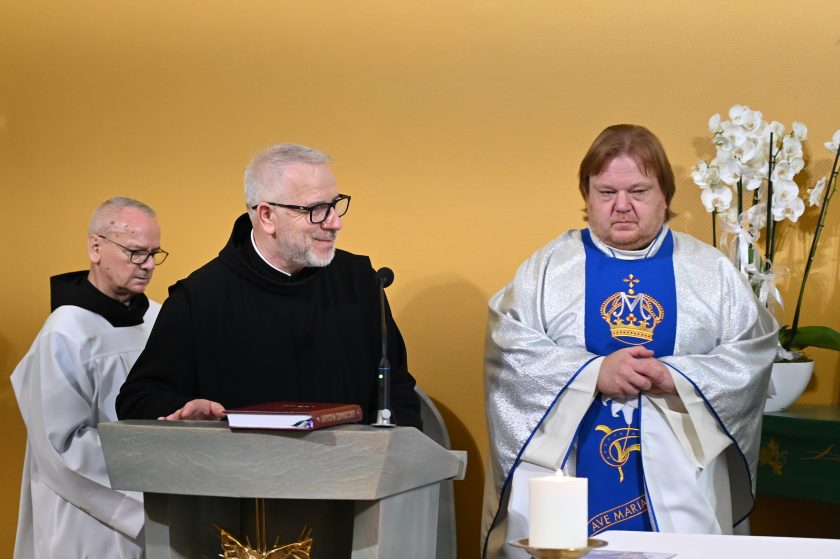 Zakonnik w czarnym habicie przemawia z ambony, uśmiechając się. Obok niego stoi ksiądz w srebrnym ornacie z niebieskim wizerunkiem maryjnym. W tle biały kwiat storczyka