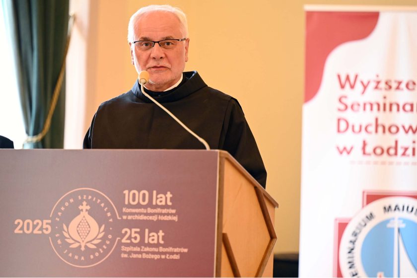 Zakonnik o siwych włosach i w okularach przemawia z mównicy z bordowym panelem i złotymi napisami jubileuszu „2025 • 100 lat… • 25 lat…”. Mikrofon na jasnym, łukowatym ramieniu skierowany ku ustom mówcy. W tle zielona kotara okienna i roll-up „Wyższe Seminarium Duchowne w Łodzi” z okrągłym znakiem graficznym.