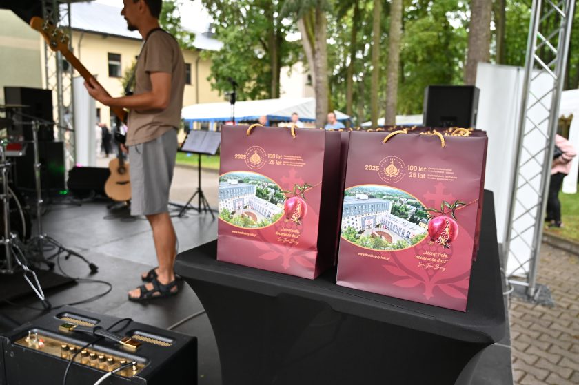 Kolorowe torby upominkowe z grafiką Szpitala Bonifratrów w Łodzi i granatem – symbolem zakonu. Torby stoją na stole, a w tle widać gitarzystę przygotowującego się do występu na scenie