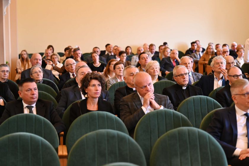 Kolejne szerokie ujęcie widowni. W pierwszym rzędzie kilka pustych miejsc, za nimi liczni uczestnicy w formalnych strojach; wśród nich osoby starsze i duchowny w koloratce. Twarze skierowane ku scenie, światło równomierne; w tle rzędy foteli zapełniają niemal całą salę.