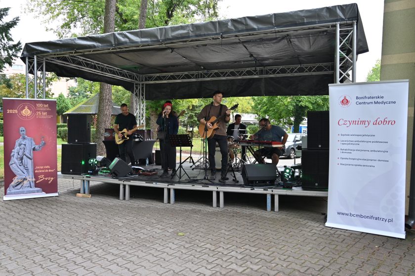 Zespół muzyczny występuje na scenie plenerowej. Wokalistka i gitarzysta grają i śpiewają, obok perkusista i klawiszowiec. Po bokach sceny banery z hasłem jubileuszowym oraz logo Bonifraterskiego Centrum Medycznego
