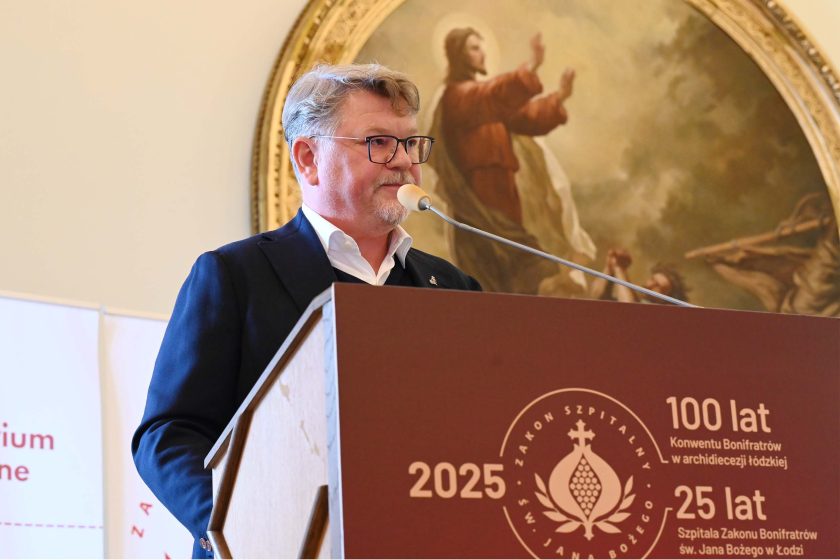Mężczyzna w okularach, w granatowej marynarce i białej koszuli przemawia z tej samej bordowej mównicy z logotypem zakonu i napisami „2025 • 100 lat… • 25 lat…”. Po lewej fragment roll-upu seminaryjnego, za mówcą duży obraz w złotej ramie z postacią w czerwonej szacie. Mikrofon na jasnym wysięgniku, na krawędzi mównicy drobne zarysowania widoczne w świetle.