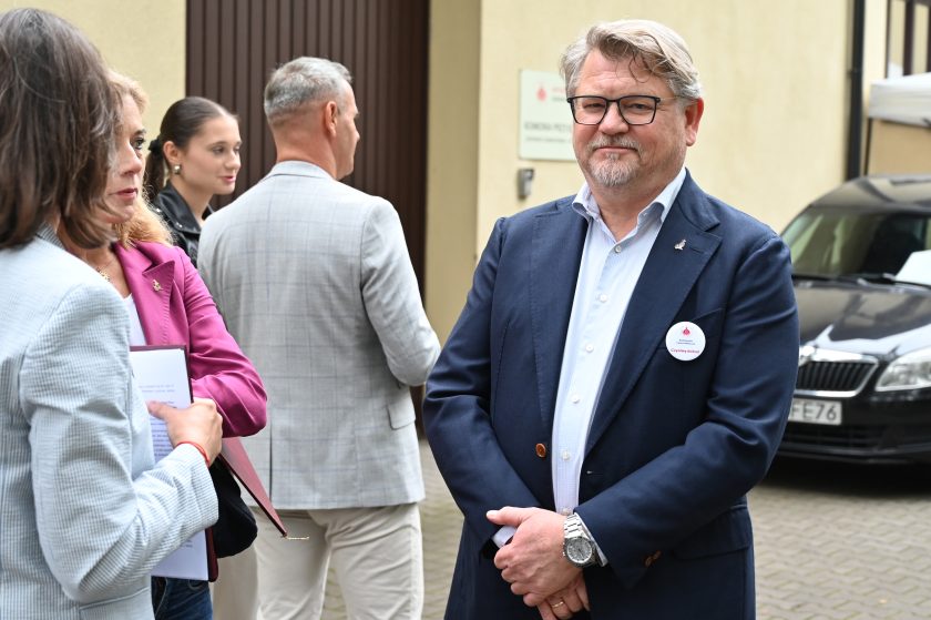 Mężczyzna w eleganckiej granatowej marynarce z przypiętym znaczkiem i identyfikatorem stoi na dziedzińcu, patrząc w stronę aparatu. W tle grupa osób prowadzi rozmowę