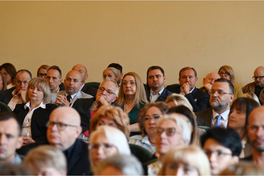 Zbliżenie środkowej części widowni w zielonych fotelach. W centrum kobieta z długimi, jasnymi włosami i okularami patrzy uważnie na scenę; obok niej mężczyźni w garniturach. Głębia ostrości płytka — pierwszy plan rozmyty, ostre twarze w środkowym rzędzie; atmosfera skupienia i ciszy.