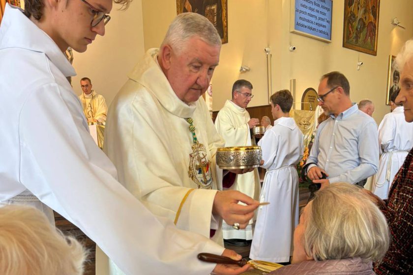 Biskup w asyście ministranta udziela komunii świętej starszej kobiecie, w tle widać duchownych i wiernych zgromadzonych przy ołtarzu