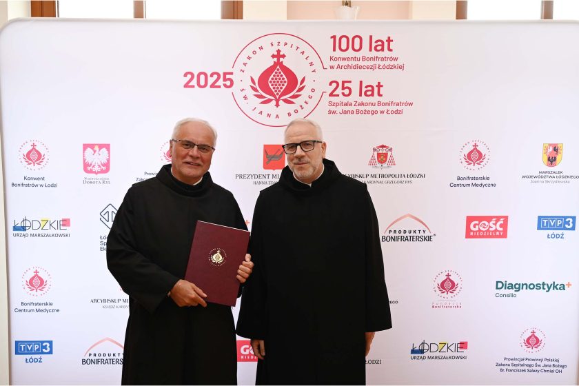 Dwóch zakonników Bonifratrów stoi razem przed ścianką jubileuszową, jeden z nich trzyma teczkę z herbem zakonu.