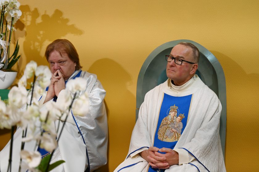 Dwaj księża w ornatach maryjnych siedzący obok siebie. Jeden w skupieniu splótł dłonie przy twarzy, drugi siedzi spokojnie z rękami złożonymi na kolanach