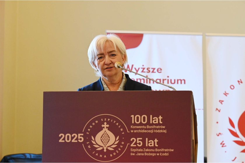 Kobieta o krótkich siwych włosach przemawia zza bordowej mównicy z emblematem w kształcie granatu z krzyżem oraz napisami „2025 • 100 lat Konwentu Bonifratrów w archidiecezji łódzkiej • 25 lat Szpitala Zakonu Bonifratrów św. Jana Bożego w Łodzi”. Ma granatową marynarkę i jasną koszulę. Z przodu mikrofon na giętym wysięgniku, w tle jasna ściana i roll-upy z logo oraz napisem „Wyższe Seminarium Duchowne w Łodzi”.