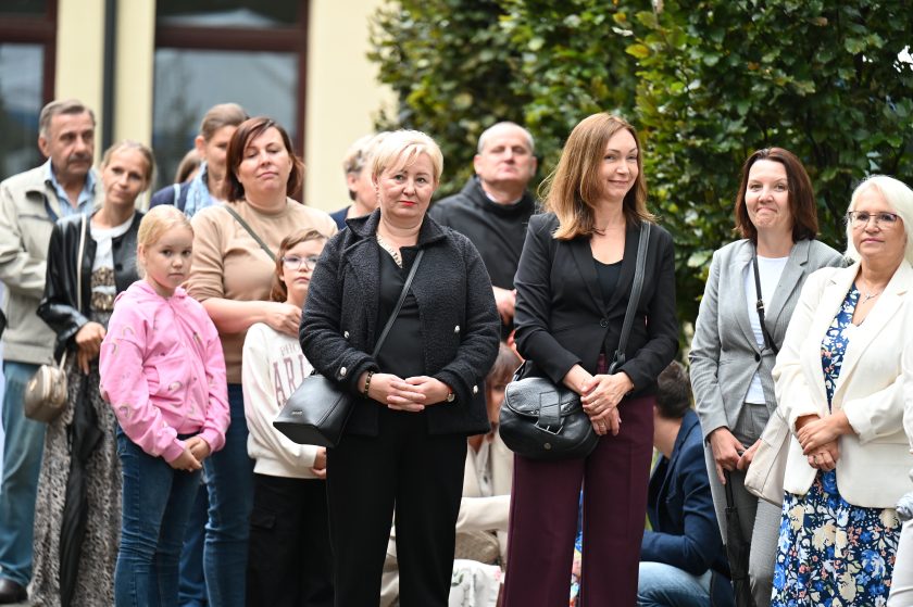 Grupa osób zgromadzonych na dziedzińcu, uśmiechnięci uczestnicy uroczystości. Na pierwszym planie dzieci i kobiety w eleganckich strojach, w tle mężczyźni i zakonnik w czarnym habicie