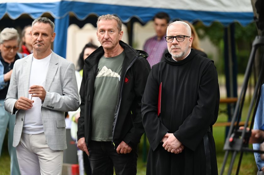 Trzech mężczyzn stojących razem podczas uroczystości. Jeden w jasnoszarej marynarce, drugi w zielonej koszulce i czarnej kurtce, trzeci – zakonnik w habicie, trzymający teczkę