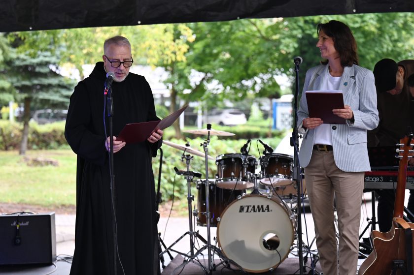 Zakonnik w czarnym habicie oraz kobieta w jasnej marynarce stoją na scenie plenerowej i czytają z teczek. W tle zestaw perkusyjny i instrumenty muzyczne