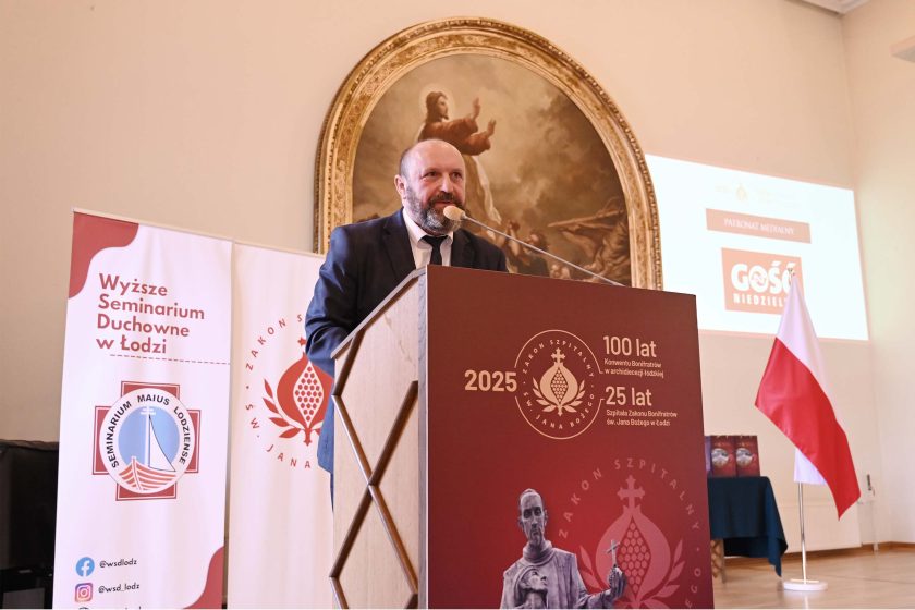 Łysy mężczyzna z brodą, w ciemnym garniturze i białej koszuli, przemawia zza tej samej bordowej mównicy. Po lewej dwa roll-upy seminaryjne, za mówcą na ścianie dominujący owalny obraz religijny, po prawej projekcja z logotypem „Gość Niedzielny” i stojak z polską flagą. Na stoliku z tyłu rząd bordowych teczek/prezentów.