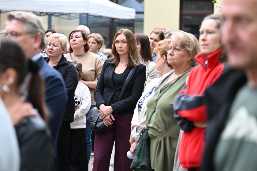 Grupa osób stojących na dziedzińcu podczas wydarzenia. Kobiety i mężczyźni słuchają uważnie, część z poważnymi minami, inne z lekkim uśmiechem