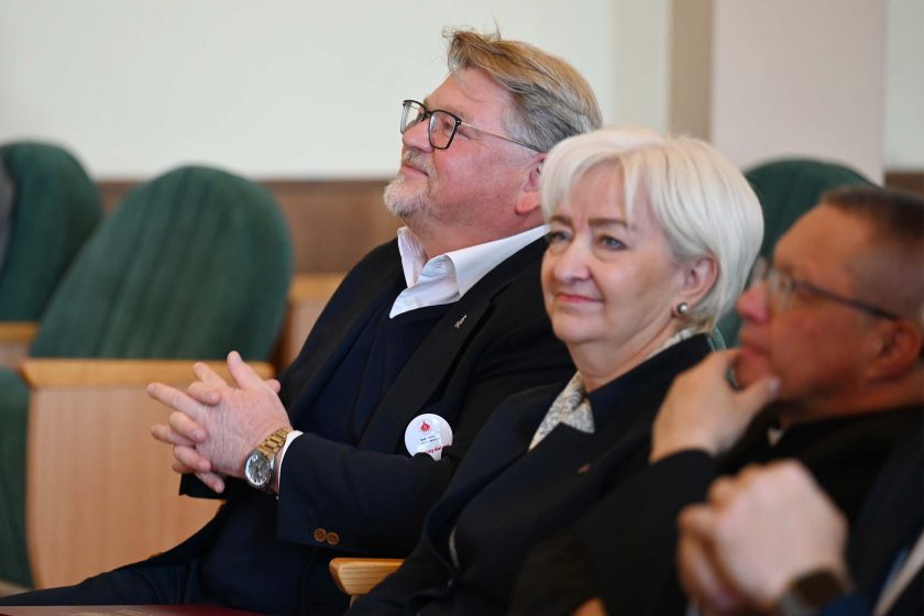 Ujęcie z widowni: mężczyzna w okularach i granatowym garniturze uśmiecha się, splatając dłonie; obok kobieta o siwych włosach lekko się uśmiecha, po prawej duchowny w sutannie z dłonią przy policzku. Zielone oparcia foteli tworzą rytmiczne tło, światło dzienne ociepla scenę.