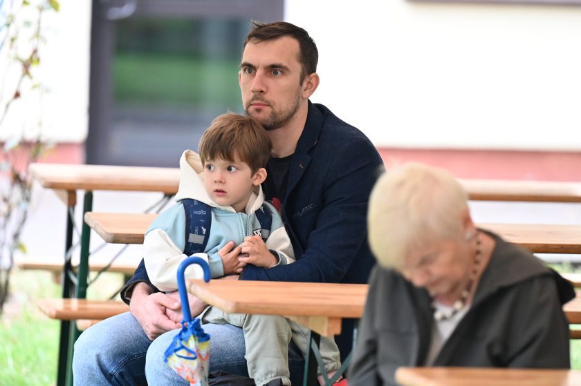 Mężczyzna siedzący na ławce plenerowej z małym chłopcem na kolanach. Dziecko w jasnej bluzie i plecaku patrzy w bok, a ojciec obejmuje je, patrząc przed siebie z zamyśleniem