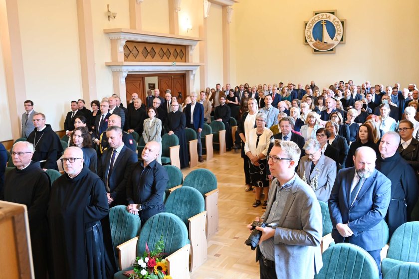 Szerokie ujęcie sali w trakcie uroczystości; większość uczestników stoi między rzędami zielonych foteli. Z przodu po lewej kilku zakonników w czarnych habitach, po prawej mężczyzna z aparatem w dłoniach. Na ścianie nad wejściem duża emblematyczna tarcza „Archidiecezja Łódzka”; w pierwszym rzędzie kompozycja kwiatowa.