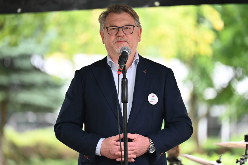 Mężczyzna w granatowej marynarce i okularach przemawia do mikrofonu na scenie plenerowej. Na piersi przypięty ma znaczek i identyfikator z napisem „Czynimy dobro”