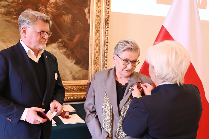 Ceremonia dekoracji: kobieta o siwych włosach przypina odznaczenie do marynarki starszej kobiety w szarym żakiecie z wzorzystą apaszką i czarnej bluzce z koronką. Obok, po lewej, mężczyzna w okularach trzyma etui na medal. Tło stanowi fragment dużego obrazu w złotej ramie, stojak z polską flagą i stół z dokumentami oraz etui dekoracyjnymi.
