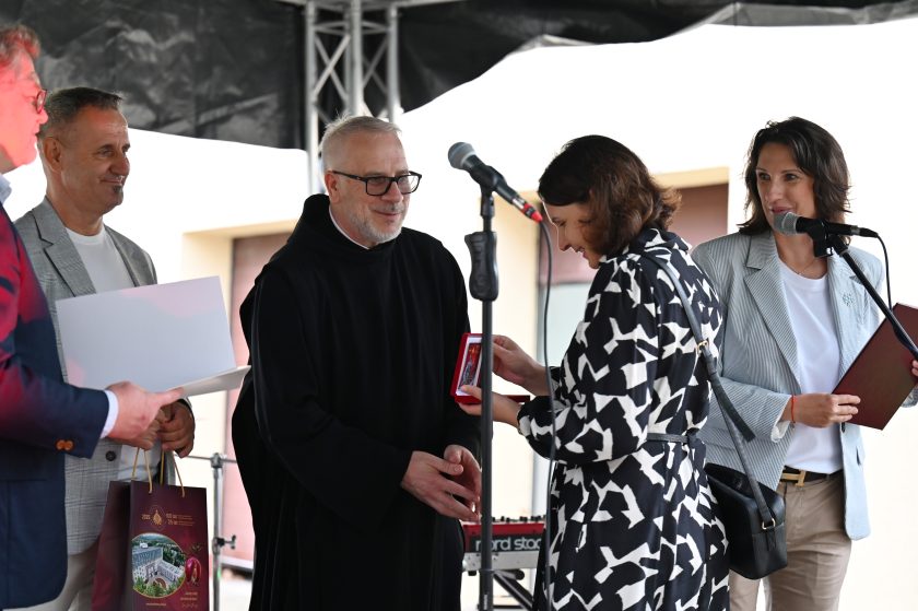 Ceremonia wręczenia nagród na scenie. Zakonnik w czarnym habicie uśmiecha się, wręczając medal kobiecie w biało-czarnej sukience. Obok kobieta w jasnej marynarce trzyma mikrofon i dokumenty