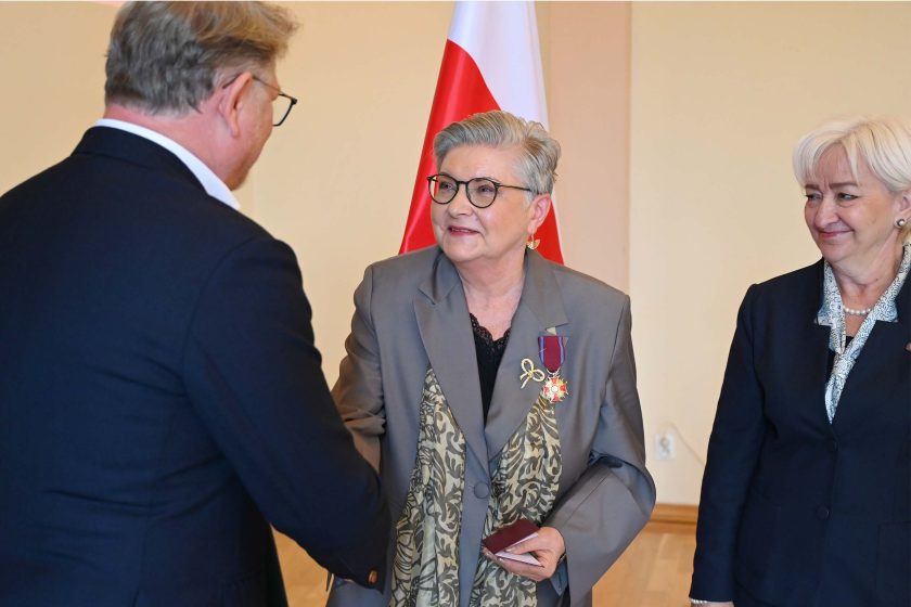 Po zakończeniu dekoracji odznaczona kobieta w szarym żakiecie ściska dłoń mężczyzny w granatowej marynarce; na jej klapie widoczne dwa odznaczenia oraz przypinka w kształcie wstążki. Obok stoi uśmiechnięta kobieta dekorująca, w granatowym kostiumie; za nimi polska flaga i jasna ściana sali.
