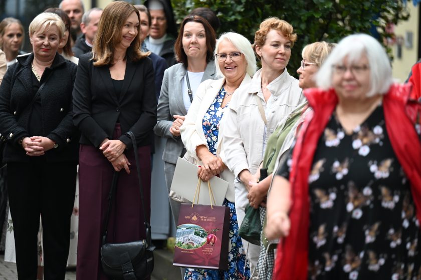 Grupa kobiet stojących razem podczas uroczystości. Kilka z nich uśmiecha się, a jedna trzyma w ręku bordową torbę upominkową z grafiką jubileuszową Bonifratrów