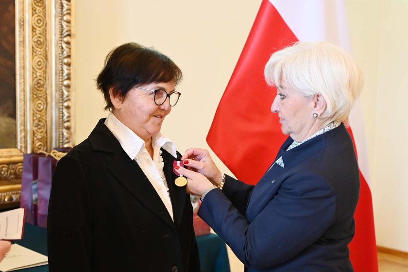 Starsza kobieta w okularach, białej bluzce i czarnym żakiecie, stoi profilowo, gdy kobieta o siwych włosach przypina jej okrągły medal z bordową wstążką. W tle polska flaga na stojaku, fragment obrazu w złotej ramie oraz stół z bordowymi torbami i etui; światło równomierne, podkreślające uroczysty charakter sceny.