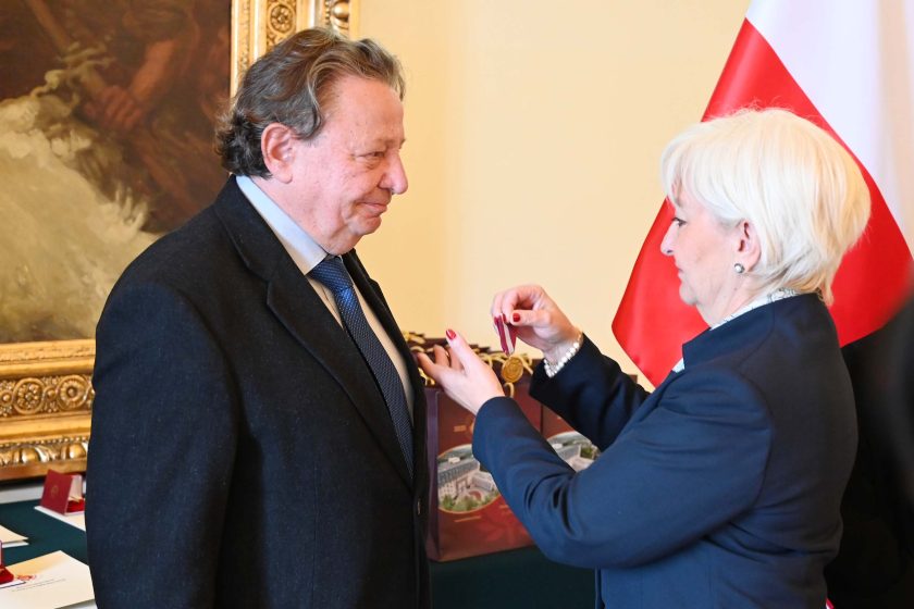 Kobieta o krótkich, siwych włosach w granatowym kostiumie przypina odznaczenie do klapy marynarki starszego mężczyzny w ciemnym garniturze i niebieskim krawacie. Na jej nadgarstku perłowa bransoletka, paznokcie pomalowane na czerwono. W tle fragment dużego obrazu w złotej, rzeźbionej ramie, po prawej stojak z polską flagą i stół z bordowymi torbami upominkowymi oraz etui na medale.