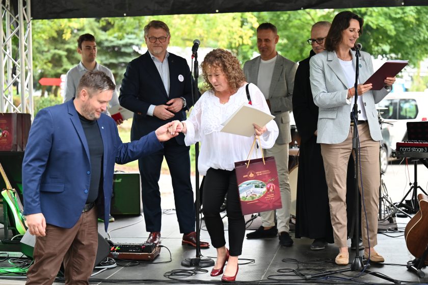 Uczestnicy wydarzenia na scenie podczas wręczenia nagród. Kobieta w białej bluzce i czarnych spodniach trzyma dyplom oraz torbę upominkową, schodząc ze sceny przy pomocy mężczyzny