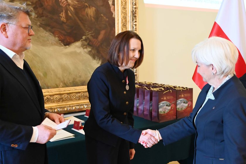 Kobieta z brązowymi włosami, z nowo przypiętym medalem na lewej piersi, pochyla się z uśmiechem i ściska dłoń kobiety dekorującej. Po lewej mężczyzna w granatowej marynarce przegląda dokumenty. Na drugim planie bordowe torby upominkowe, złota rama obrazu i biało-czerwony maszt flagowy.